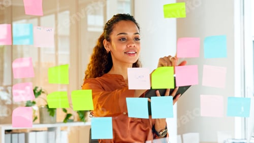 Preview: Happy female designer planning ideas on a glass wall with colorful sticky notes inside a creative a