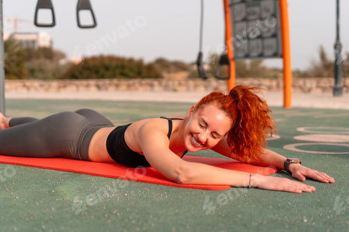 Preview: Smiling redhead woman lying on yoga mat at summer park