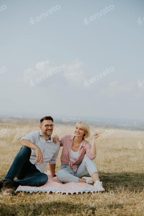 Preview: Couple enjoys a sunny day on a picnic blanket in a grassy field with scenic views