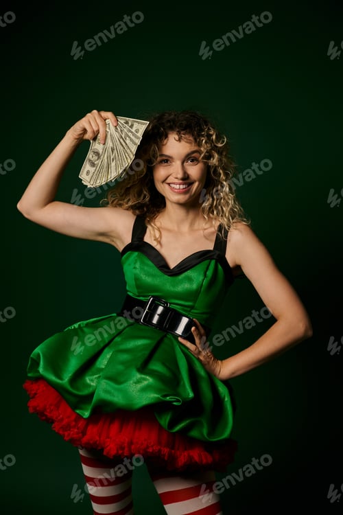 Preview: happy curly woman dressed as new year elf with one arm akimbo and with money in another hand