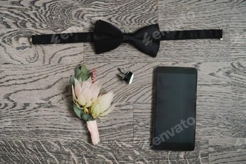 Preview: Top view of modern groom's outfit: bow tie, smart phone and boutonniere on wooden background.