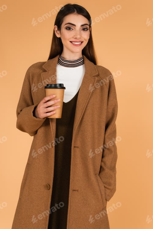 Preview: happy woman with coffee to go standing with hand in pocket of stylish coat isolated on beige