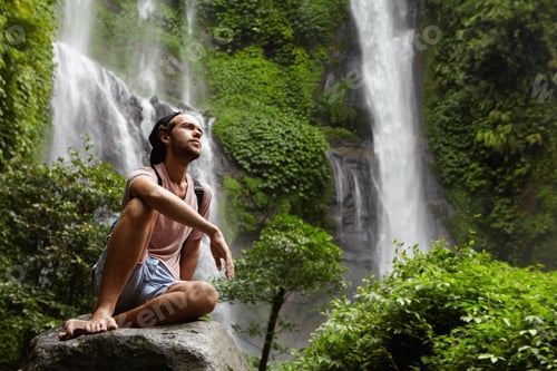 Preview: Attractive young bearded adventurer wearing no shoes having break on big rock while trekking alone i