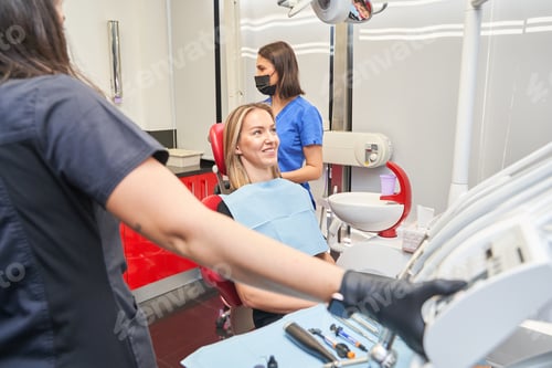 Preview: dentist with face mask talking to blonde woman during dental procedure at dental clinic.