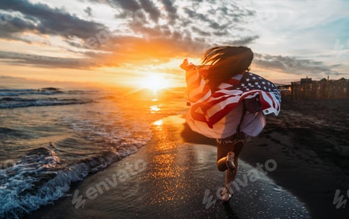 Preview: Young woman holding national American flag walking ocean beach. America Independence Day concept