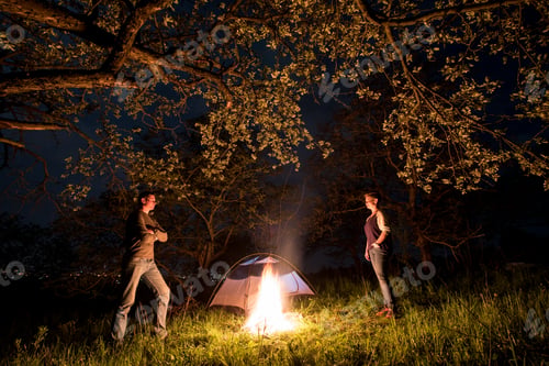 Preview: Couple tourists standing at a campfire near tent under trees and night sky. Night camping