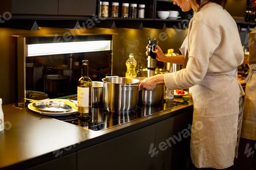 Preview: Woman in linen apron cooking with blender pumpkin soup in saucepan on induction oven in dark kitchen