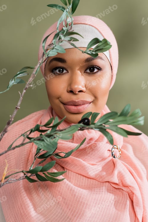 Preview: portrait of young multiracial woman in pink hijab smiling at camera near olive branch on green