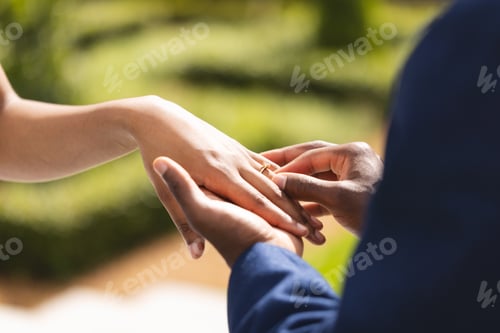 Preview: Hands of african american bride and groom placing ring on finger at wedding ceremony in sunny garden