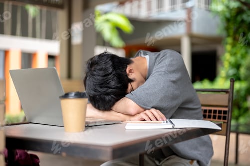 Preview: Glasses asian man napping sleeping on arms in front of laptop and book while sitting at wooden table