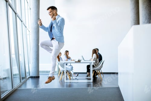 Preview: Young businessman with smartphone jumping in a modern office