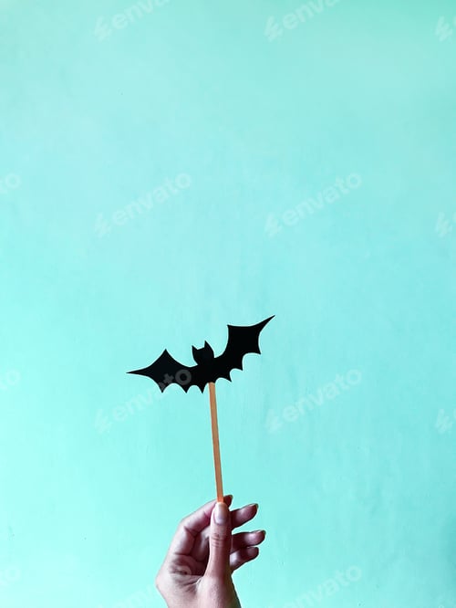 Preview: Female Hand Holding Black Paper Bat In Front Of Blue Monochrome Background For Halloween Party Fun