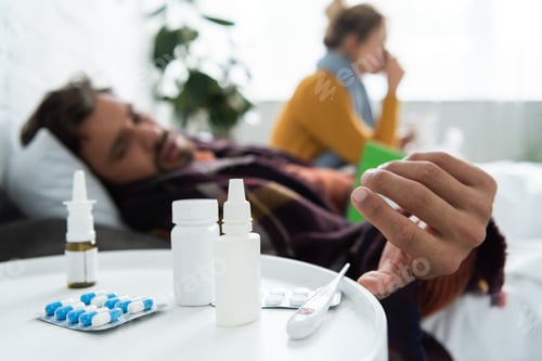 Preview: selective focus of sick lying in bed with thermometer, hot drink and nasal spray and pills on table