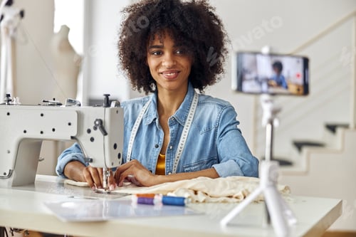Preview: African-American seamstress sews clothes with modern machine shooting video lesson in workshop
