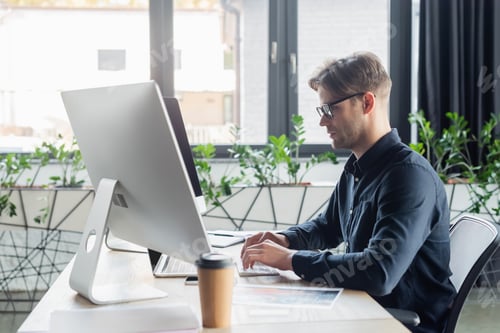Preview: Side view of programmer using computers near blurred document and coffee in office