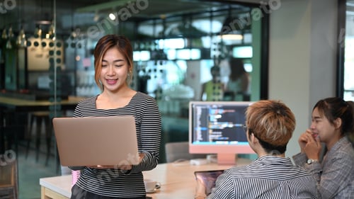 Preview: Asian female team leader standing in office and software developers team working in background.