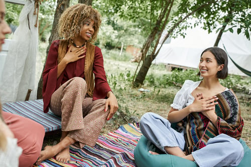 Preview: smiling multiethnic women in boho outfits meditating on meadow outdoors in retreat center