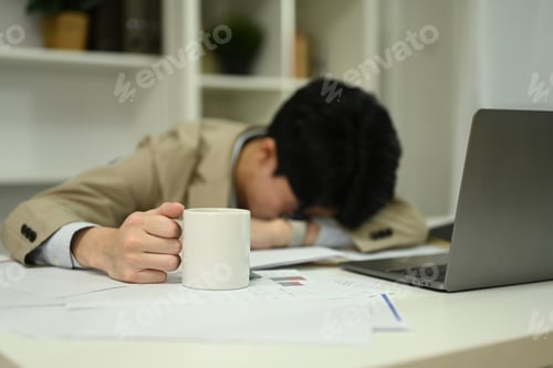 Preview: Tired office worker laying down on desk with one hand holding a coffee mug.