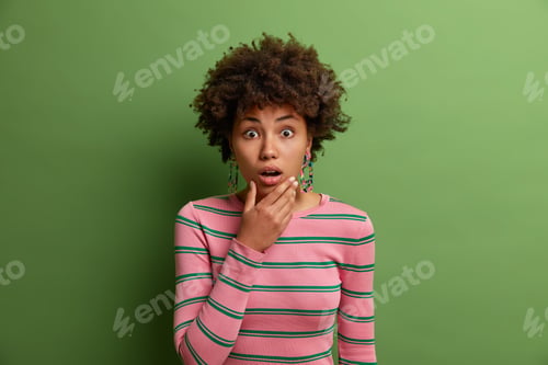 Preview: Shocked speechless ethnic curly woman holds chin and looks with bugged eyes at camera, amazed and st