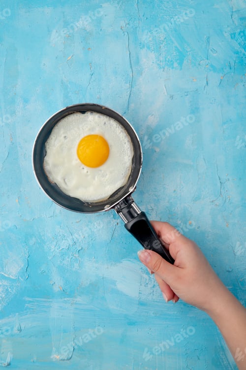 Preview: top view of woman hand holding frying pan with fried egg on blue background with copy space