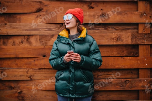 Preview: Portrait of woman in emerald green coat on wooden background. Girl in red hat and sunglasses holdin