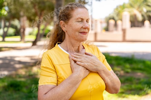 Preview: Senior woman standing in park with eyes closed, hands on chest, feeling gratitude and peace