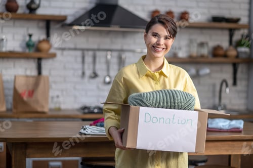 Preview: Cheerful female volunteer demonstrating a cardboard box full of clothes