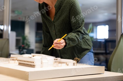 Preview: Midsection of caucasian female architect analyzing model on desk with pencil at modern workplace