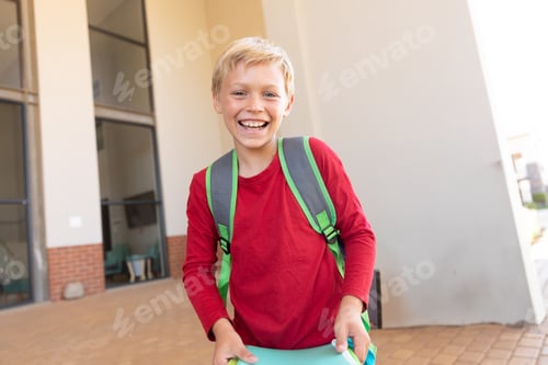 Visualização: Menino sorridente com mochila segurando livro fora do prédio da escola, sentindo-se animado