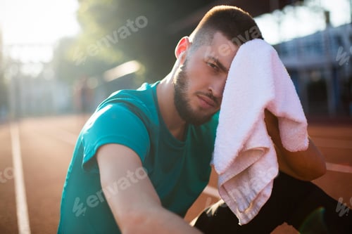 Preview: Young sporty man tiredly looking in camera while holding white towel in hand on racetrack of stadium