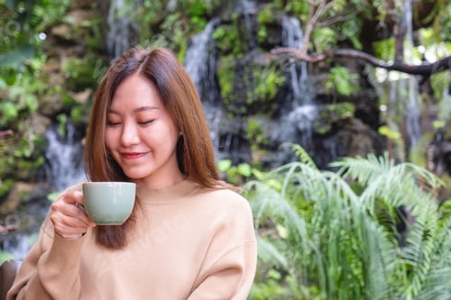 Preview: A young woman holding and drinking coffee while sitting in the garden with waterfall