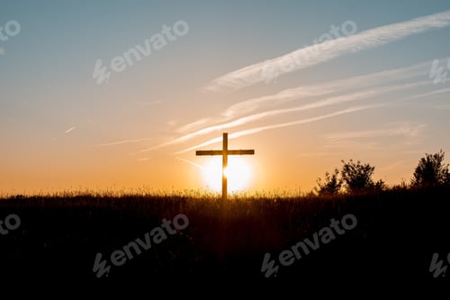 Preview: Silhouette of a cross in a grassy field with the sun shining in the background