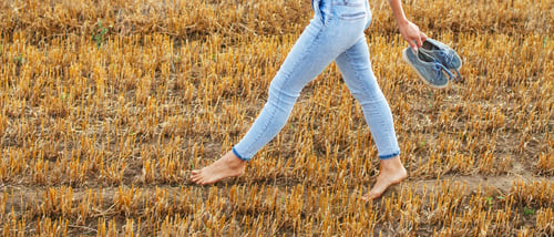 Preview: barefoot girl with sneakers in hand walking in the agricultural field with haystack and bales