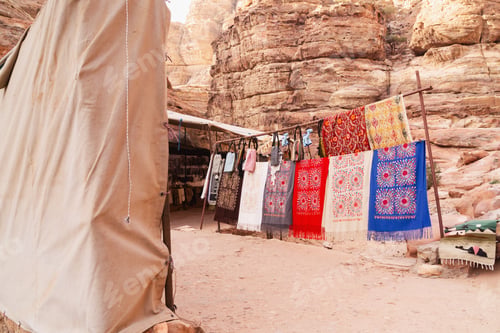 Preview: Colorful fabrics hang on display near rock formations in a warm desert location. Petra, Jordan.