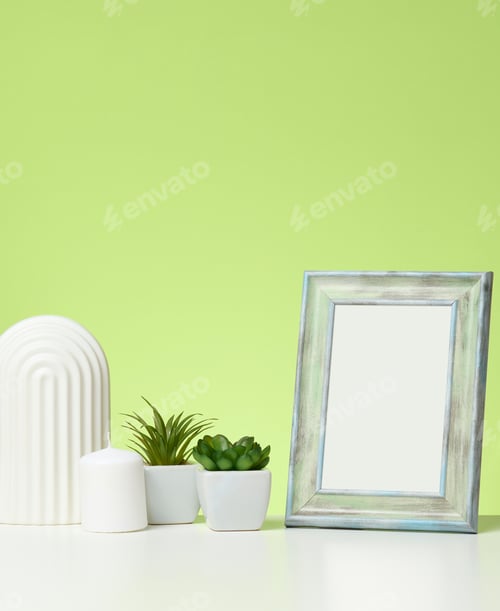 Preview: Empty white wooden photo frame and flowerpots with plants on white table, green background