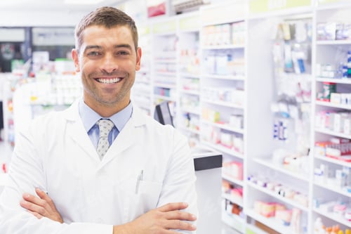 Preview: Young adult male pharmacist standing behind counter in pharmacy with medicine shelves, copy space