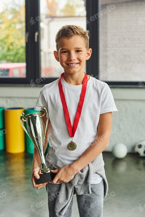Preview: vertical shot of little cute boy with golden medal and trophy smiling at camera, child sport