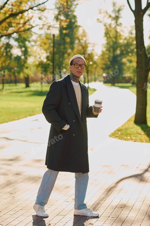 Preview: Stylish young man with tattoos enjoys a coffee stroll in a sunlit urban park setting