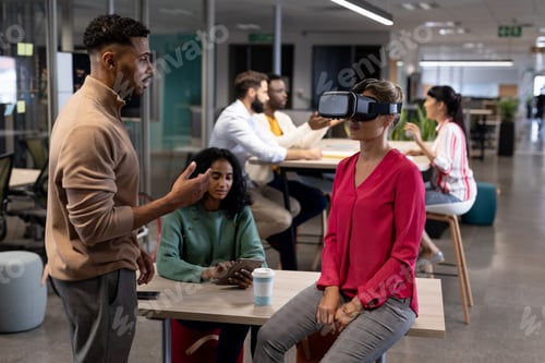 Preview: Hispanic businesswoman discussing with caucasian businesswoman wearing vr simulator in meeting