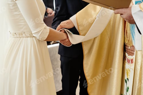 Preview: priest holding hands of stylish bride and elegant groom at catholic wedding ceremony