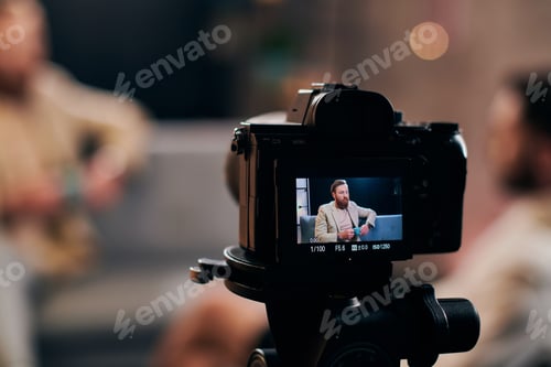Preview: focus on camera filming stylish blurred man with beard in elegant attire discussing questions