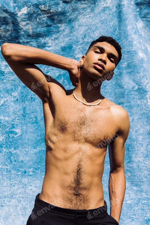 Preview: young shirtless african american man in beads posing with hand behind neck near blue drape