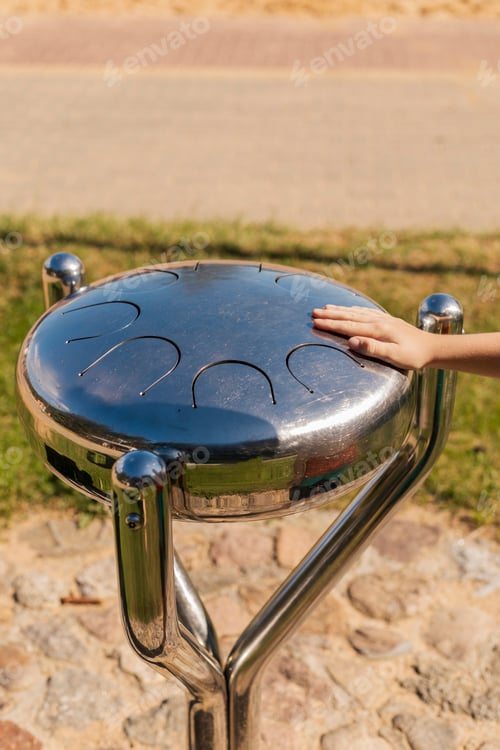 Preview: A child's hand gently rests on a shiny metal musical instrument in a park.