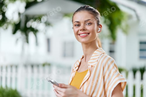 Preview: Smiling woman using smartphone outdoors with wireless earbuds, wearing a striped shirt, in a lush