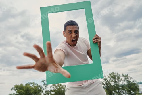 Preview: Playful mixed race man stretching out hand from picture frame with sky in the background