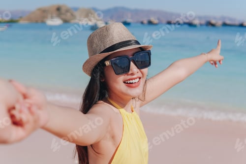 Preview: Cheerful young Asian woman holding hands on Pink Beach, Labuan Bajo while pointing to the scenery.