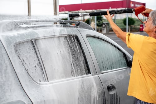Preview: Smiling caucasian senior woman washing her car in a self-service car wash station using high pressur