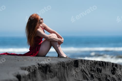 Preview: Woman in red long dress with raised hem and bare legs sitting on black sand on beach, hugging knees