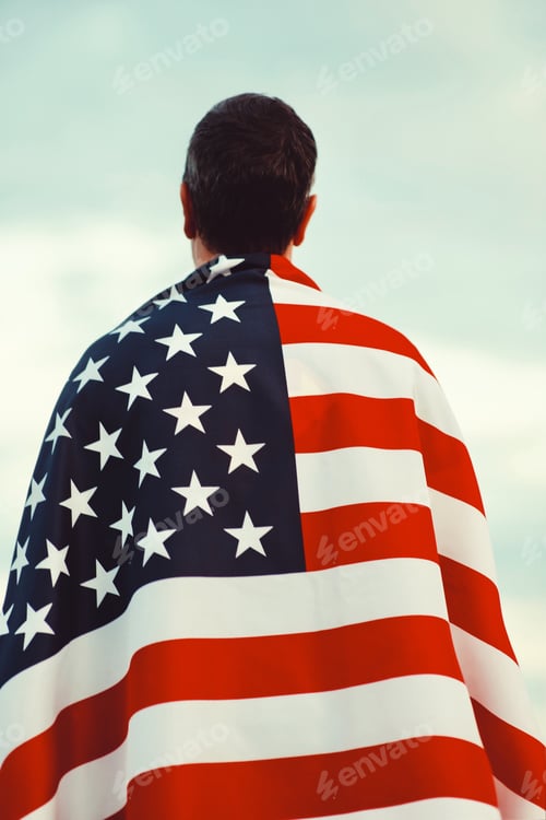 Preview: Back view millennial candid authentic man wrapped in US flag looks at the sky on celebrate July 4th