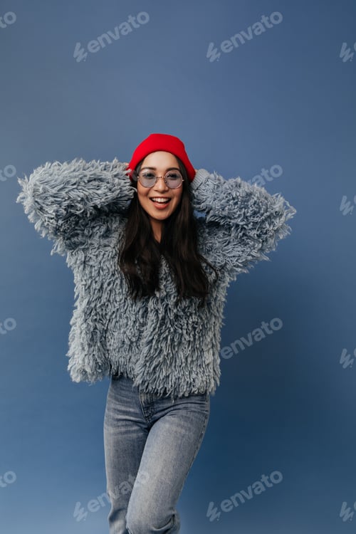 Preview: Positive brunette lady with red cap and round blue sunglasses in stylish fluffy sweater and jeans l